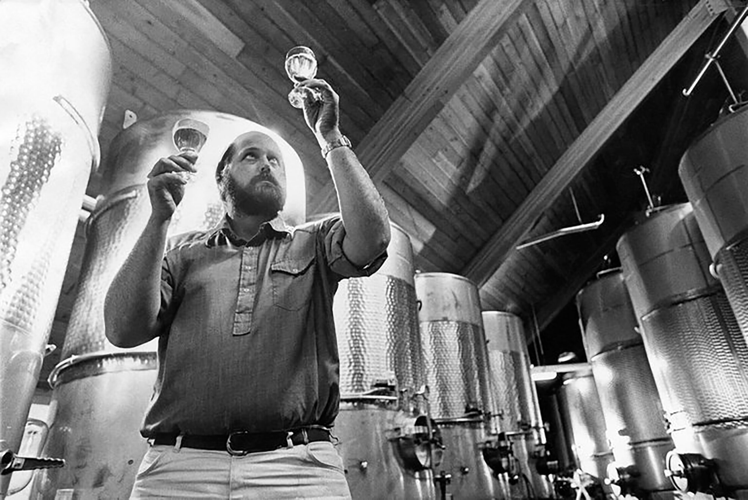 Don Chappellet inspecting two glasses of wine next to silver tanks